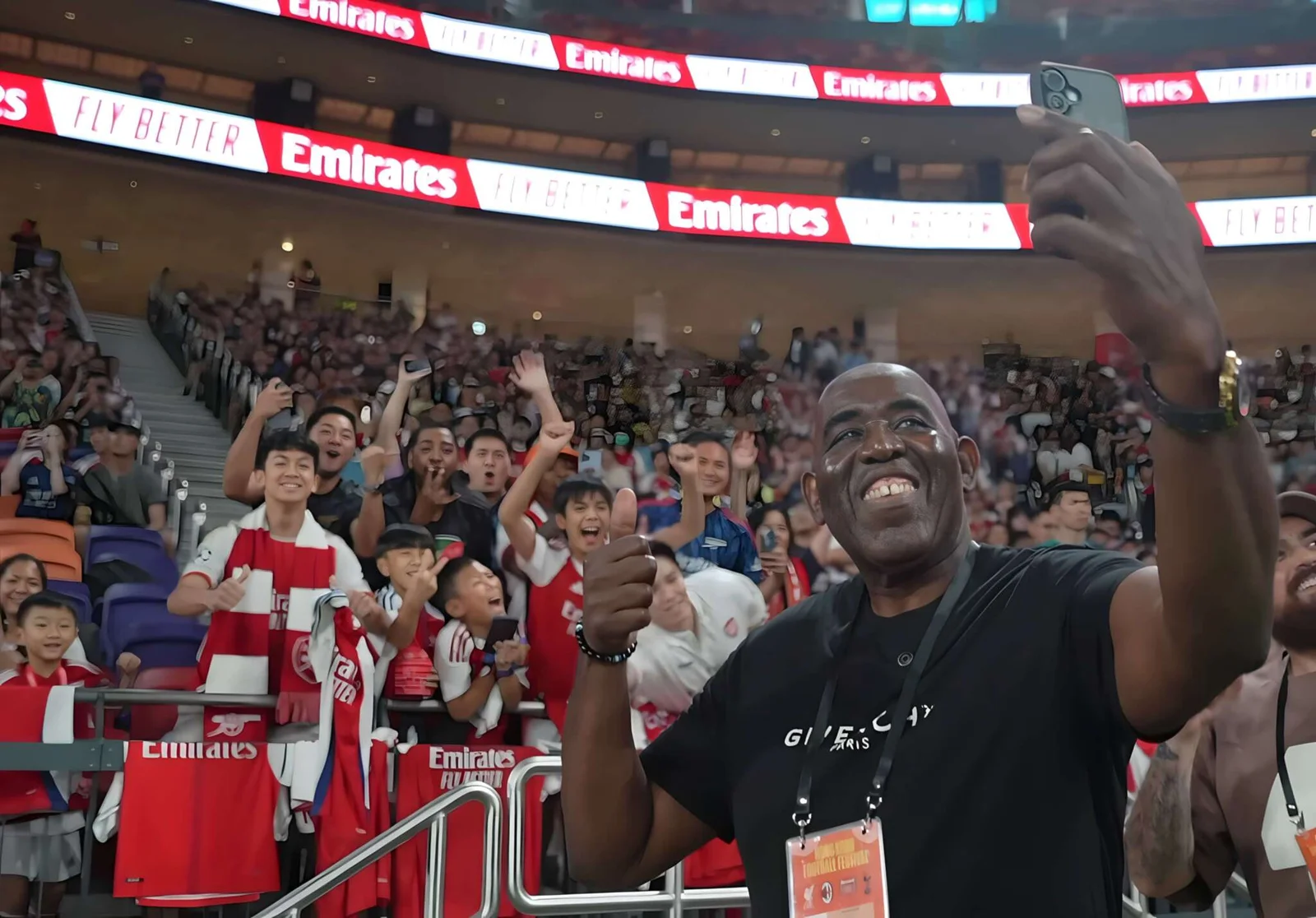GFN — a smiling fan takes a selfie in a packed stadium with supporters cheering behind him.
